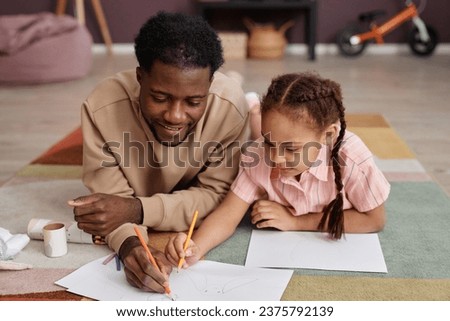 Portrait of Black father and daughter drawing pictures together laying on floor at home and smiling