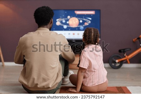 Back view of father and daughter watching educational videos about space together sitting on floor in front TV