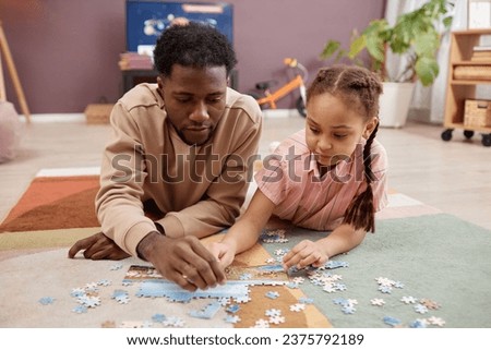 Front view portrait of father and daughter solving jigsaw puzzle together while lying on soft carpet at home enjoying family time