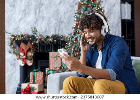 Happy smiling christmas man using phone watching funny video smiling and listening to online music, streaming podcasts, hispanic man using headphones sitting on sofa for new year holidays.