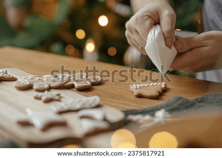 Hands decorating gingerbread cookies with icing on rustic wooden table at christmas tree golden lights. Atmospheric Christmas holiday traditions, family time