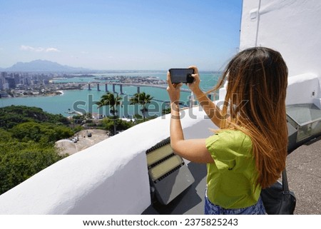 Young traveler woman takes picture with her smartphone of Vitoria city from Penha Convent in Vila Velha, Espirito Santo, Brazil