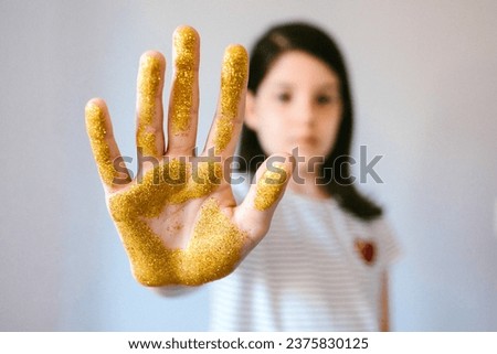 Serious young girl showing her palm hand covered of golden glitter while doing stop sign. Concept of harmful microplastics composition to humans and environment. Selective focus on hand. Royalty-Free Stock Photo #2375830125