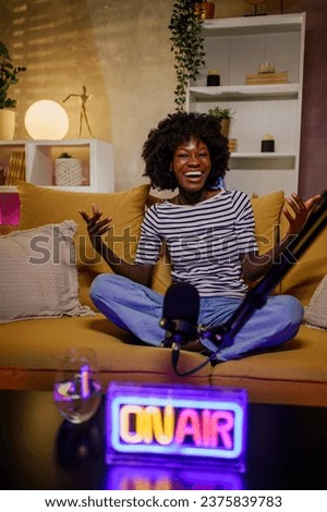 Portrait of an african american smiling female radio host recording a new internet podcast episode while sitting on a yellow sofa near microphones in cozy broadcasting studio. Podcast concept.