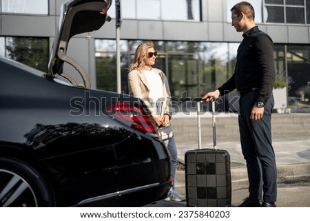 Chauffeur packs a suitcase in a car trunk, businesswoman waiting nearby using luxury taxi service during a business trip. Concept of business transfer services, idea of personal driver. Royalty-Free Stock Photo #2375840203