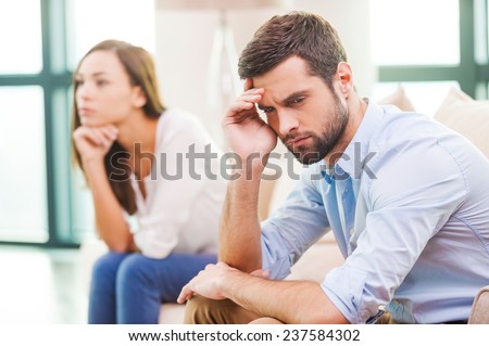 Relationship breakdown. Depressed young man holding hand on head and looking away while woman sitting behind him on the couch  Royalty-Free Stock Photo #237584302