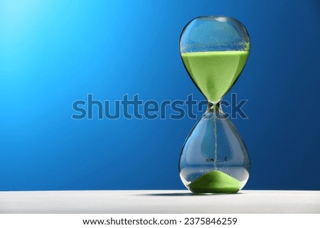Hourglass with light green flowing sand on white table against blue background. Space for text Royalty-Free Stock Photo #2375846259