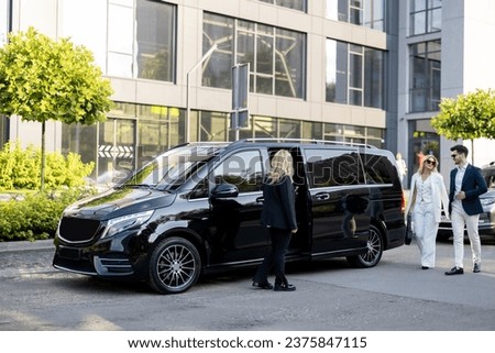 Female chauffeur waits a business people to let them in a minivan taxi, keeping door open. Concept of business trips and transportation service Royalty-Free Stock Photo #2375847115