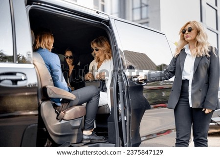 Female chauffeur helps a business lady to get out of vehicle, opening a door of luxury black minivan. Concept of personal driver, luxury taxi for business delegation Royalty-Free Stock Photo #2375847129