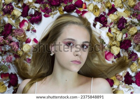 Cute blonde girl with makeup lies in the petals and buds of wilted flowers and looks at the camera