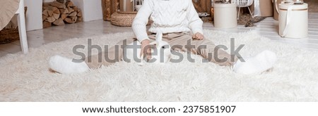 Christmas concept. child with white rabbit on the white carpet near Christmas tree in a home interior. Symbol of the year 2023. Close-up. Chinese New year.