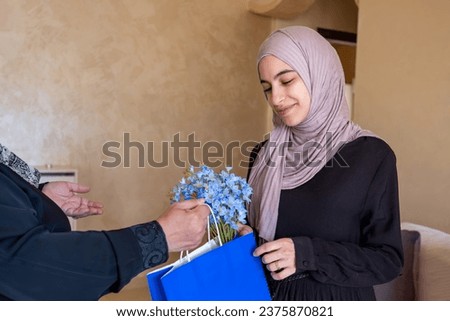 Arabic woman giving present or gift to her mother wearing hijab and abaya inside living room for mother day with blue roses