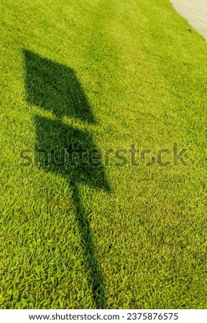 Shadow of a street sign up close on a large field on bright green grass on a sunny day. The shadow shows the long pole holding the sign and two signs.