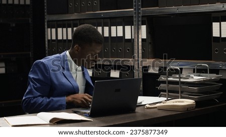 Determined private detective analyzing confidential documents from shelves in an investigation. African american investigator cross-checks evidence and statements on personal computer and case files.