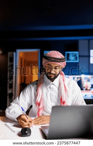 Professional Muslim guy uses technology to work efficiently in the office, browsing online and writing notes from a laptop. Youthful Arab male student using his notebook and minicomputer for research.