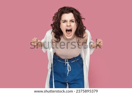 Portrait of angry woman with curly hair wearing casual style outfit standing arguing with somebody, clenched fists, screaming with hate and aggression. Indoor studio shot isolated on pink background. Royalty-Free Stock Photo #2375895379