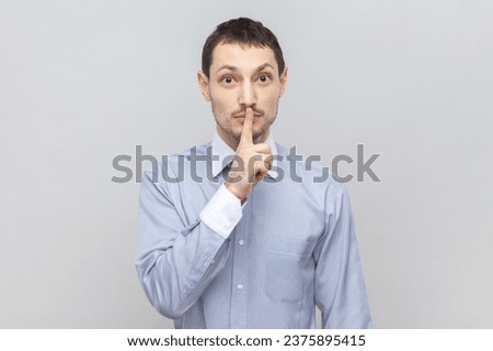 Portrait of serious concentrated man standing with finger near lips, showing shh gesture, asking to keep silence during lesson, wearing light blue shirt. Indoor studio shot isolated on gray background
