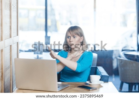 Portrait of strict serious young woman with blonde hair in blue shirt working on laptop, having video call, showing x sign gesture, no way. Indoor shot in cafe with big window on background.