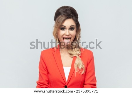 Portrait of funny crazy attractive blonde woman standing sticking tongue out, demonstrates childish behavior, wearing red jacket. Indoor studio shot isolated on gray background. Royalty-Free Stock Photo #2375895791