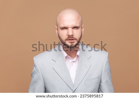 Portrait of aggressive bald bearded man looks grumpy, feels offended and displease, clenched teeth, looking at camera, wearing gray jacket. Indoor studio shot isolated on brown background. Royalty-Free Stock Photo #2375895817