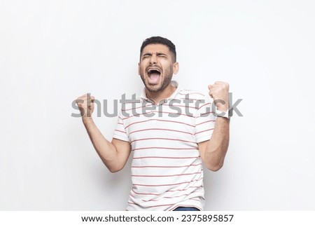 Portrait of extremely happy joyful cheerful bearded man wearing striped t-shirt standing with clenched raised fists, screaming, celebrating victory. Indoor studio shot isolated on gray background. Royalty-Free Stock Photo #2375895857