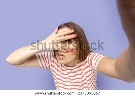 Portrait of spying woman in T-shirt taking selfie picture, point of view, looking at the camera through her finger, peeping, making front selfportrait. Indoor studio shot isolated on purple background