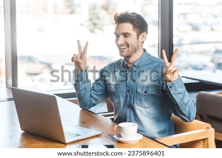 Portrait of smiling satisfied happy man freelancer in blue jeans shirt working on laptop, has video call, showing v sign to scree, rejoices his victory. Indoor shot near big window, cafe background.