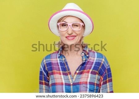 Cheerful playful mature woman in shirt, hat and eyeglasses, being in good mood, smiling broadly and winking at camera, feelings, attitude and reaction. Indoor studio shot isolated on yellow background Royalty-Free Stock Photo #2375896423