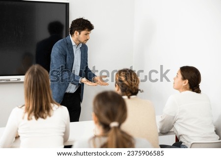 Male business coach standing near interactive board explaining topic while standing against group of people sitting at desks in front of him