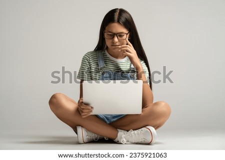 Interested student teen girl in glasses watching lection class on laptop on gray background. Focused teenager busy learning studies, study online. Education in college high school university concept. Royalty-Free Stock Photo #2375921063