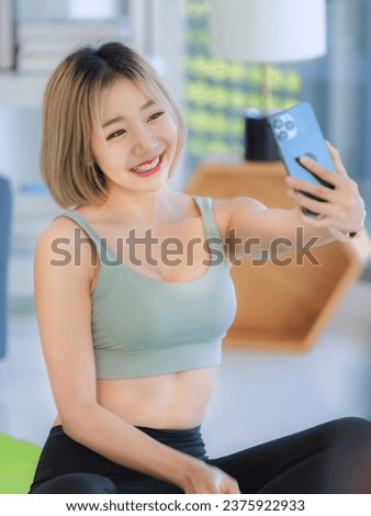 Asian young fit female athlete teenager in sportswear sport bra sitting smiling on yoga pilates mat holding smartphone in hand taking selfie photo in gym after training exercising working out.