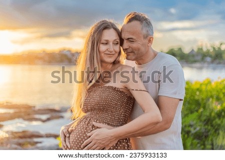 A happy, mature couple over 40, enjoying a leisurely walk on the waterfront On the Sunset, their joy evident as they embrace the journey of pregnancy later in life Royalty-Free Stock Photo #2375933133