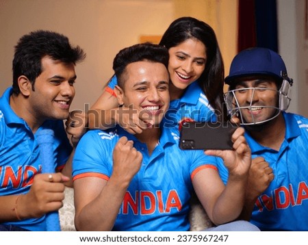 Thrilled friends in Indian jerseys cheering for team India during cricket match. Excited cricket fans sitting at home watching games on mobile, using smartphone apps to online bet