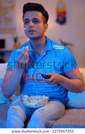 Random shot of a man having popcorn while watching a cricket match on television - wearing a jersey. A young man watching television wearing an Indian cricket team jersey at home - eating popcorn
