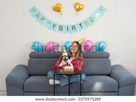 Joyful teenage girl celebrating her dog's birthday with tasty cake, wearing festive hats at home. Adorable Jack Russell Terrier having b-day party with own.  Cake for a dog.