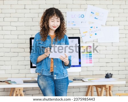 Asian professional successful young female creative graphic designer in casual fashionable denim jeans jacket standing smiling holding colors chart working in workspace in company office.