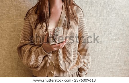 An unrecognizable brunette girl in a beige sweater holds a Christmas gift in eco packaging in her hands. a small gift in honor of Christmas, new year.