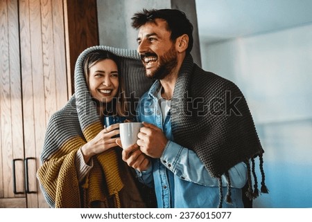 Happy young couple with hot drinks cuddling under warm blanket at home Royalty-Free Stock Photo #2376015077