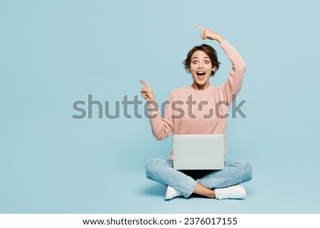 Full body young IT woman wear beige knitted sweater casual clothes sit hold use work on laptop pc computer point finger aside on area isolated on plain pastel blue cyan background. Lifestyle concept