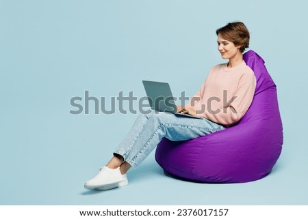 Full body young fun IT woman she wear beige knitted sweater casual clothes sit in bag chair hold use work on laptop pc computer isolated on plain pastel light blue cyan background. Lifestyle concept