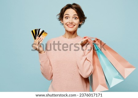 Young surprised woman wears beige knitted sweater casual clothes hold credit bank cards paper package bags after shopping isolated on plain pastel blue background. Black Friday sale buy day concept