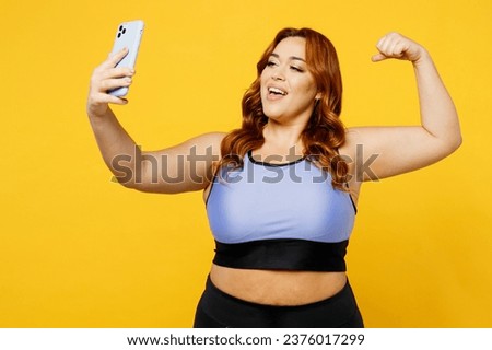 Young chubby overweight plus size big fat fit woman wear blue top warm up train do selfie shot mobile cell phone show muscles isolated on plain yellow background studio home gym. Workout sport concept