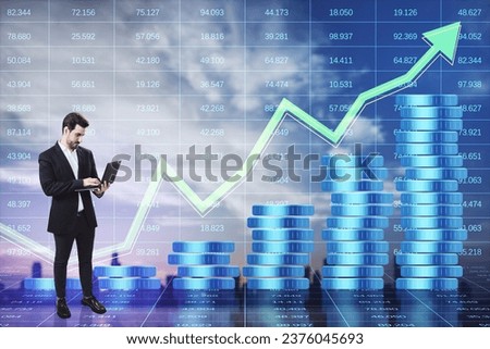 Thoughtful young businessman with laptop and growing blue coins stack hologram on blurry blue index city background with arrow. Saving money concept. Double exposure