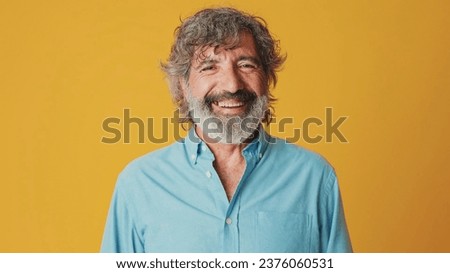 Close-up of an elderly grey-haired bearded man wearing a blue shirt, looking at the camera and laughing isolated on an orange background in the studio Royalty-Free Stock Photo #2376060531