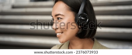 Close up portrait of smiling asian girl in headphones, listens to music outdoors, looking happy. People concept
