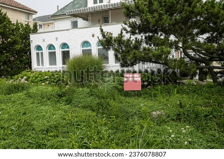A red sign in a overgrown yard in front of a large stucco house that says, "stop Windmills. Save our Shores"