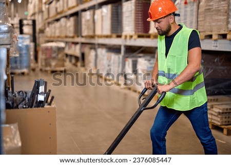 male staff worker in warehouse uses hand pallet stacker to transport goods, alone, dressed in working clothes and safety hard hat.Skilled warehouse employee pushing manual pallet jack Royalty-Free Stock Photo #2376085447