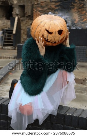Halloween costume. a girl in a lush green fur coat and a blue-violet dress and orange tights with a pumpkin on her head with slits for the eyes and mouth. ruins in the background