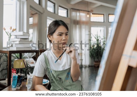 Female artist thinking and hold paintbrush while looking at canvas and pondering about paintin