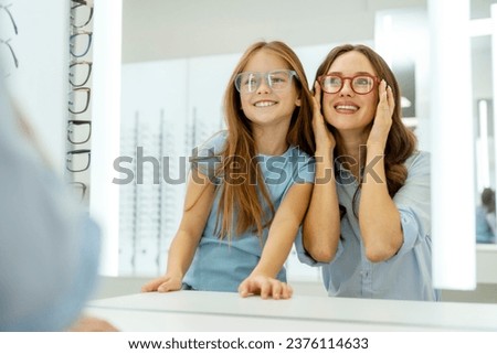 Beautiful smiling mother and daughter choosing eyeglasses looking at mirror in optical store. Advertisement, health care, vision concept  Royalty-Free Stock Photo #2376114633